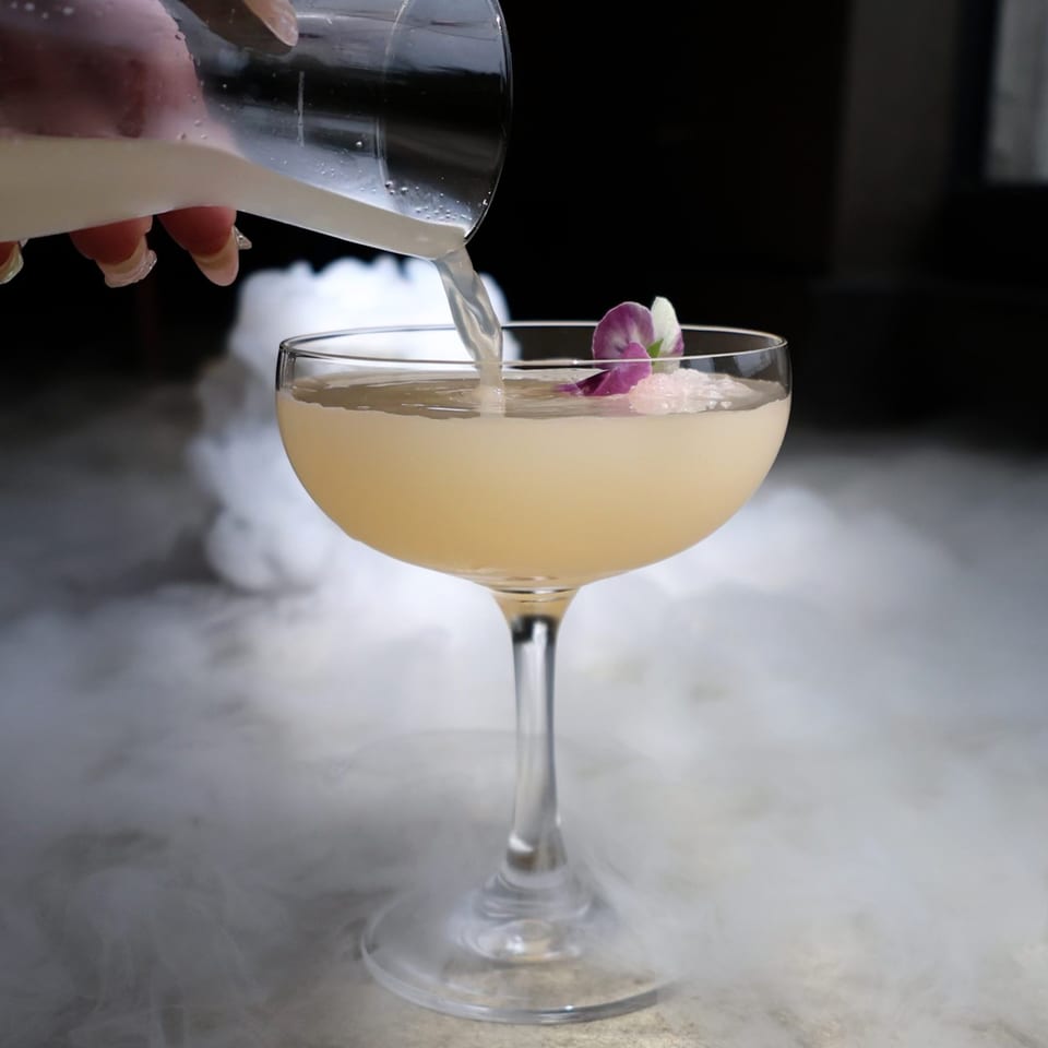A hand pours a light-colored cocktail into a glass with a purple orchid, surrounded by mist.