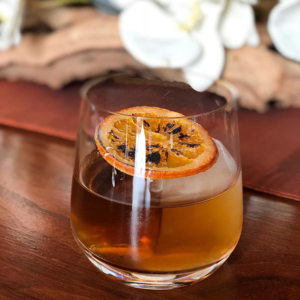An AnQi Bistro cocktail with a charred orange slice and ice on a wooden table.
