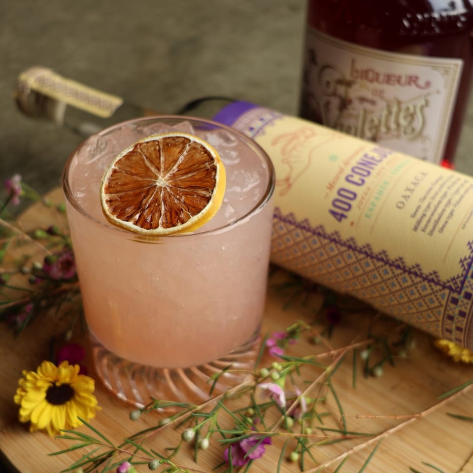 Pink cocktail with a dried citrus slice, surrounded by flowers and liquor bottles on a wooden surface.
